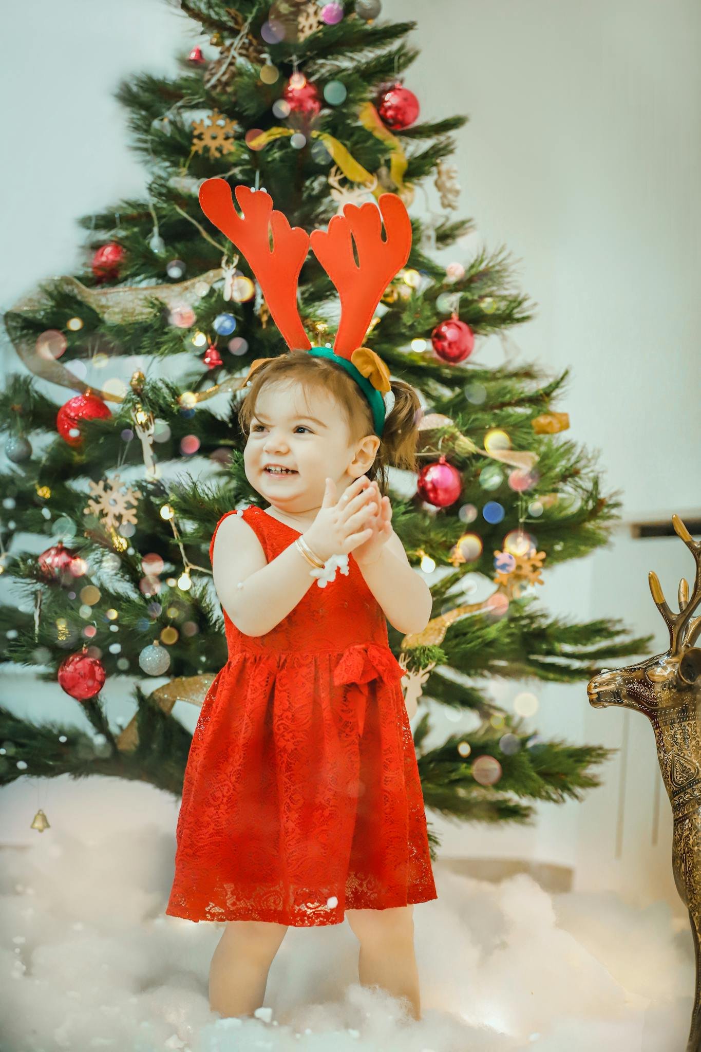 Cute child in festive outfit with antlers at Christmas tree, smiling.