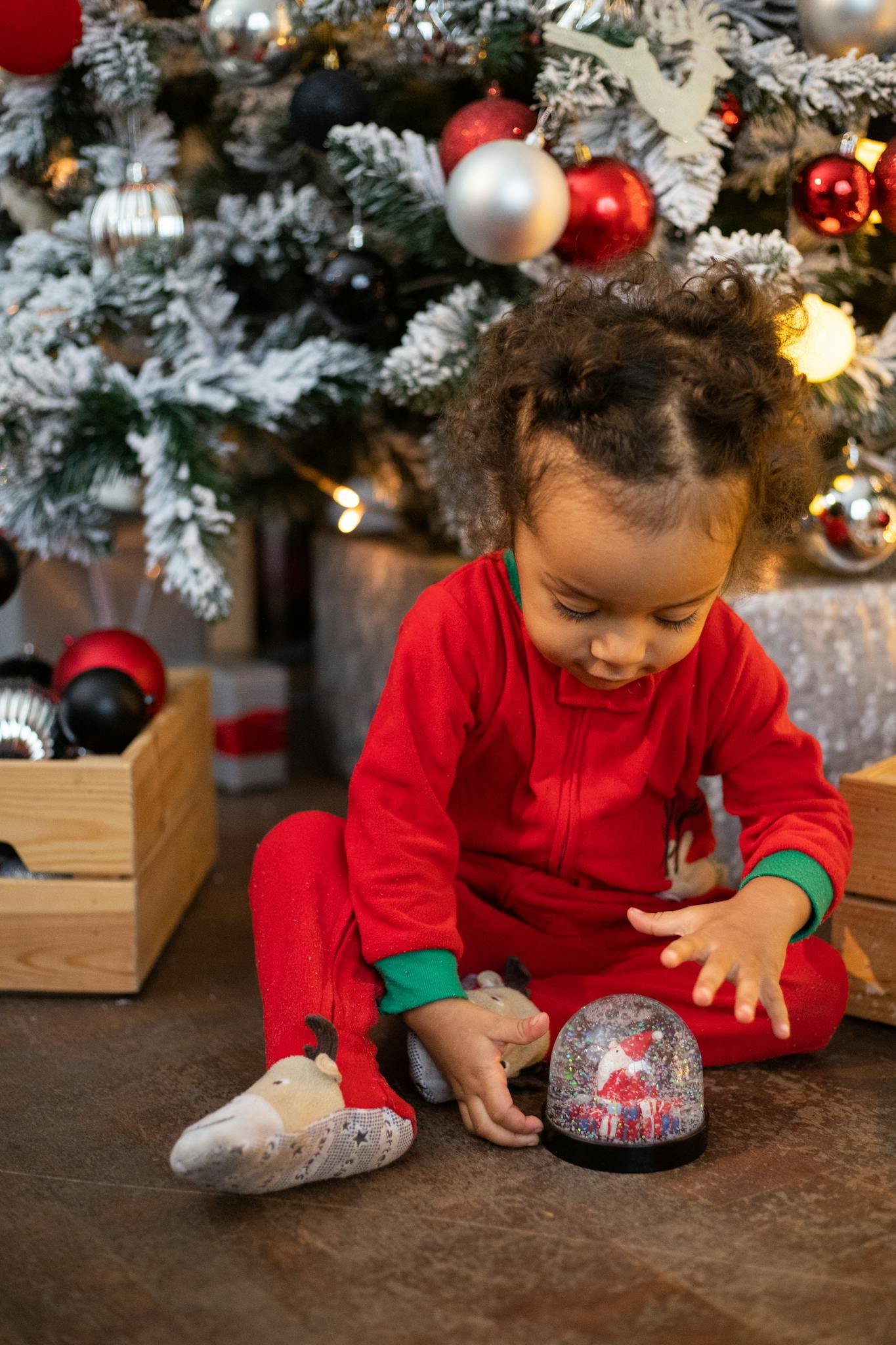 Young child in red pajamas plays with a snow globe under a Christmas tree, capturing holiday joy.